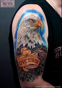 an eagle tattoo on the arm with live to ride written in gold and ribbon around it