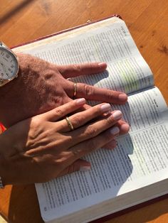 two people holding hands over an open book on a wooden table with a clock in the background