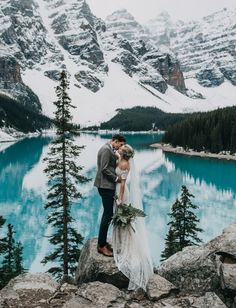 a bride and groom standing on the edge of a cliff overlooking a lake with snow covered mountains in the background