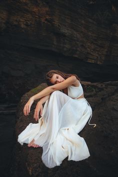 a woman sitting on top of a rock next to a river wearing a white dress