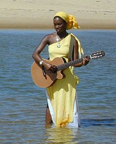 a woman standing in the water holding a guitar and wearing a yellow dress with a bandana on her head