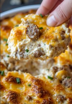 a hand holding a piece of food over a casserole dish with meatballs and cheese