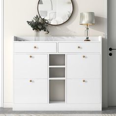 a white cabinet with two drawers and a round mirror on the wall above it, next to a lamp