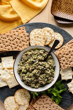 a black plate topped with crackers and a white bowl filled with green dip surrounded by crackers