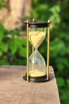 an hourglass sitting on top of a wooden table in front of some trees and bushes