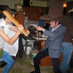 a man holding a large long loaf of bread in front of a woman sitting at a table