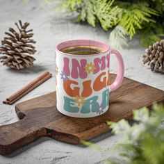 a pink and white coffee mug sitting on top of a wooden board next to pine cones