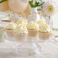 three cupcakes with white frosting on a clear cake plate and flowers in the background