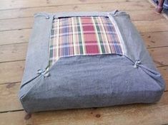 a grey ottoman with plaid fabric on the bottom and ties at the top, sitting on a wooden floor