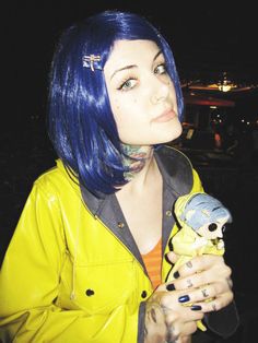 a woman with blue hair and piercings holding a stuffed animal in her right hand