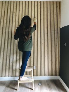 a woman standing on a ladder painting a wall with wood slats in the background