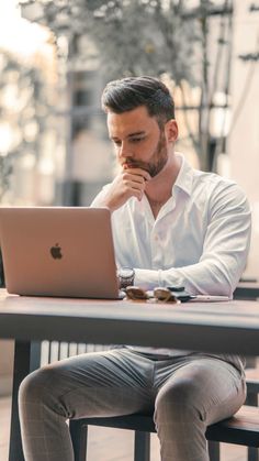 a man sitting at a table looking at his laptop computer while holding his chin to his face