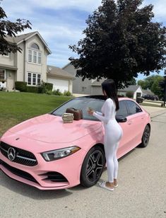 a woman standing next to a pink car in front of a house with her purse on the hood