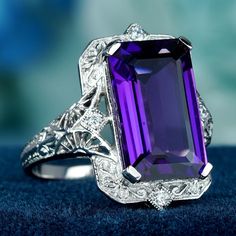 The ring is indeed a showstopper! The mesmerizing 5.8 carat emerald-cut amethyst, with a deep purple hue is both luxurious and eye-catching. The prong setting allows the amethyst to take center stage, while the intricate filigree detailing in a floral pattern on the band adds a touch of vintage charm. The diamonds set around the amethyst add a touch of sparkle and contrast, enhancing the ring's brilliance and elevating its luxury appeal.  CHARACTERISTICS Status: Made to order Origin: Thailand Me Luxury Oval Amethyst Ring In Art Deco Style, Luxury Amethyst Art Deco Rings, Luxury Art Deco Purple Amethyst Ring, Art Deco Amethyst Hallmarked Rings, Luxury Hallmarked Art Deco Amethyst Ring, Purple Amethyst Art Deco Jewelry, Vintage Amethyst Filigree Jewelry, Amethyst Color, Purple Hues