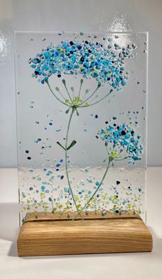 a glass vase with blue flowers in it on a wooden base and white surface behind it