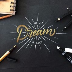 we all dream in gold written on a black paper with some pens and pencils next to it