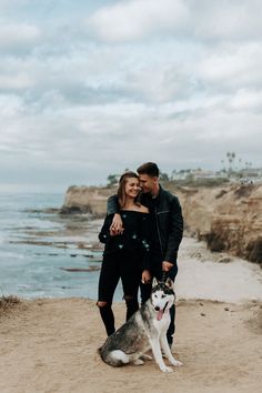 a man and woman standing next to a dog on the beach with water in the background