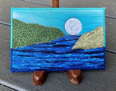 a painting on a wooden easel with a clock in the sky above water and mountains