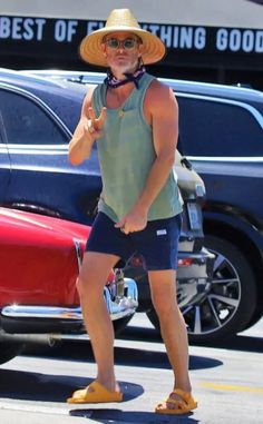 a man with a hat and sunglasses giving the thumbs up sign in front of cars