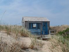 a small blue building sitting on top of a sandy beach next to grass and bushes