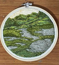 an embroidery project with mountains and trees in the background on a wooden surface, hanging from a hook