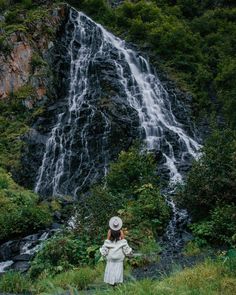 a woman standing in front of a waterfall wearing a white dress and hat with her hands on her hips