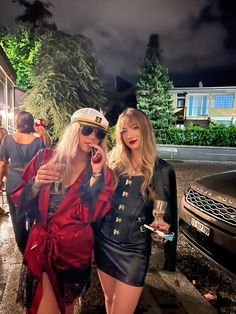 two beautiful women standing next to each other in front of a black car at night