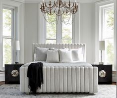 a white bed sitting under a chandelier in a bedroom next to two windows