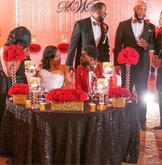 a group of people standing around a table with red flowers and candles in front of them