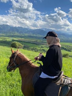 a woman riding on the back of a brown horse through a lush green field with mountains in the background