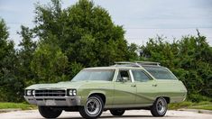 an old green station wagon parked in a parking lot next to some trees and bushes