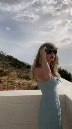 a woman in sunglasses is standing on a ledge and talking on her cell phone while looking off into the distance