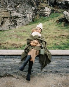 a woman holding a baby wrapped in a blanket on top of a stone wall next to a mountain