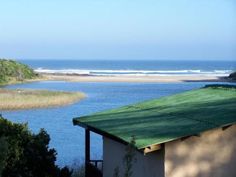 a house with a green roof next to the ocean and trees in front of it