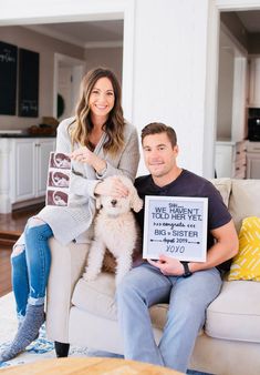 a man and woman sitting on a couch holding a sign with a dog in front of them