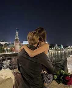 a man and woman are hugging in front of the water at night with lights on