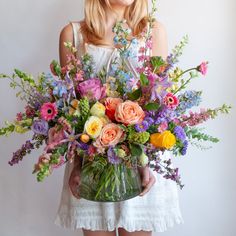 a woman in a white dress holds a glass vase with a large flower arrangement in it. The flowers are many colors and include roses, tulips, daisies, snapdragon, clematis, stock, hyacinth, and delphinium. Wildflowers Arrangements, Wild Flowers For Wedding, Wild Flower Centerpieces, Whimsical Floral Arrangements, Colorful Flower Arrangements, Colorful Floral Arrangement, Colorful Floral Wedding, Wild Flower Wedding, Wild Flower Arrangements