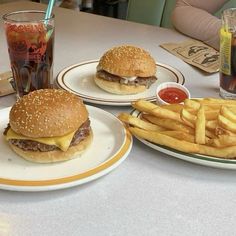 two plates with hamburgers and french fries next to a glass of soda on a table