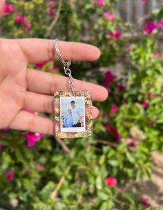 a hand holding a small square photo keychain in front of some pink flowers