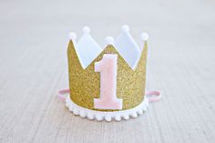 "* QUICK SHIP 3-5 days MINI CROWN: It is a mini crown in sparkly gold with white felt liner, decorated with a pink and white 1, trimmed with mini white pom poms along bottom and on crown points. Attached to a pink 1/8\" elastic- to be worn comfortably like a headband. Diameter of crown is 2.75\", and height 2.25\". *Note: Last picture is just to show example of size on a 1 year old. That crown can be found in my shop. CAKE TOPPER: Gold glitter \"one\" attached to a twine with pink/white poms on First Birthday Crown, Mini Crown, Birthday Gold, Girl 1st Birthday, Gold And Pink, My Son Birthday, Birthday Crown, Glitter Gold, Gold Birthday