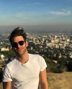 a man wearing sunglasses standing on top of a hill next to a cityscape