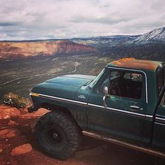 a green truck parked on top of a rocky hill with mountains in the back ground