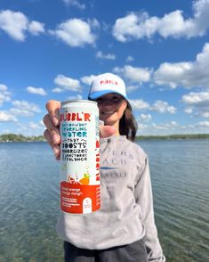 a woman holding up a can of water in front of a body of water on a sunny day