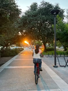 a woman riding a bike down a street next to a lamp post and trees with the sun setting in the background