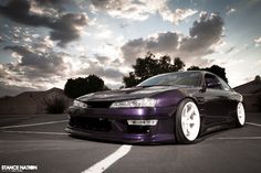 a purple car parked in a parking lot with the sun shining through clouds behind it