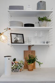 a kitchen shelf filled with dishes and plants on top of a white counter next to a wooden cutting board