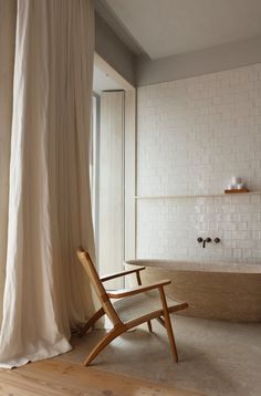 a wooden chair sitting in front of a bathtub next to a window with white curtains