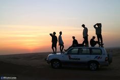 a group of people standing on top of a white truck in the desert at sunset