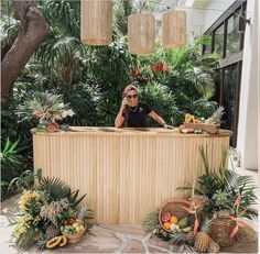 a woman standing behind a wooden counter surrounded by tropical plants and fruit in front of her