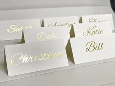 place cards with gold lettering on them sitting on a white table top next to a vase